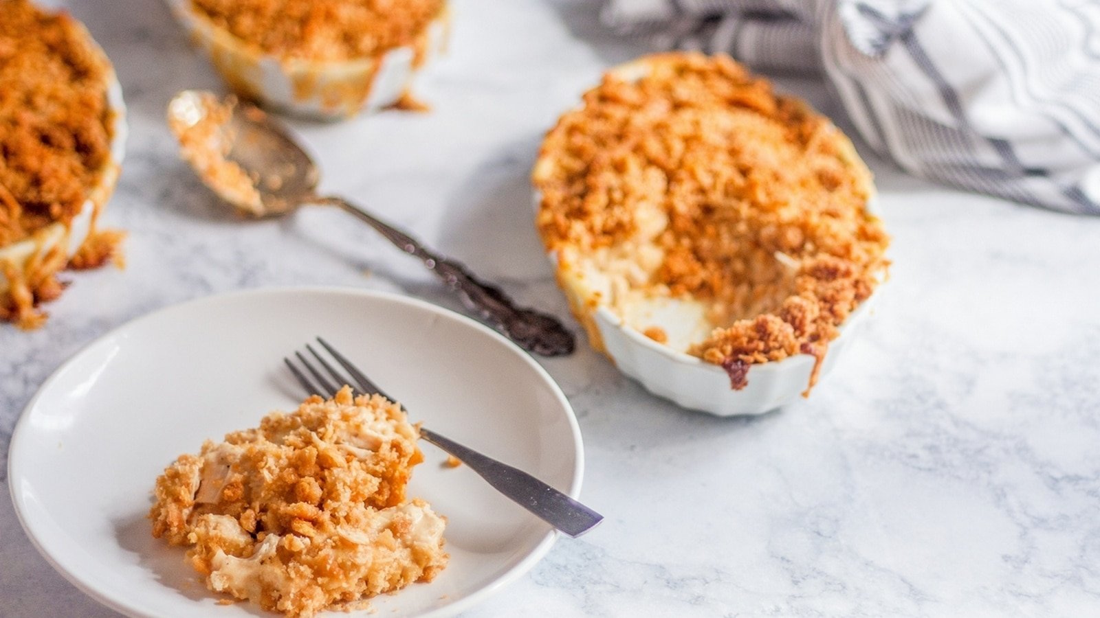 A serving of creamy macaroni and cheese with a breadcrumb topping is on a white plate with a fork. Additional portions are in the background on a marble surface.