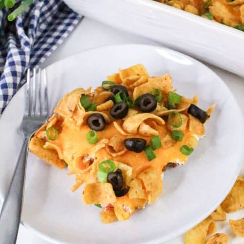 A plate of casserole topped with melted cheese, Fritos, sliced black olives, and chopped green onions. A fork is placed on the side.