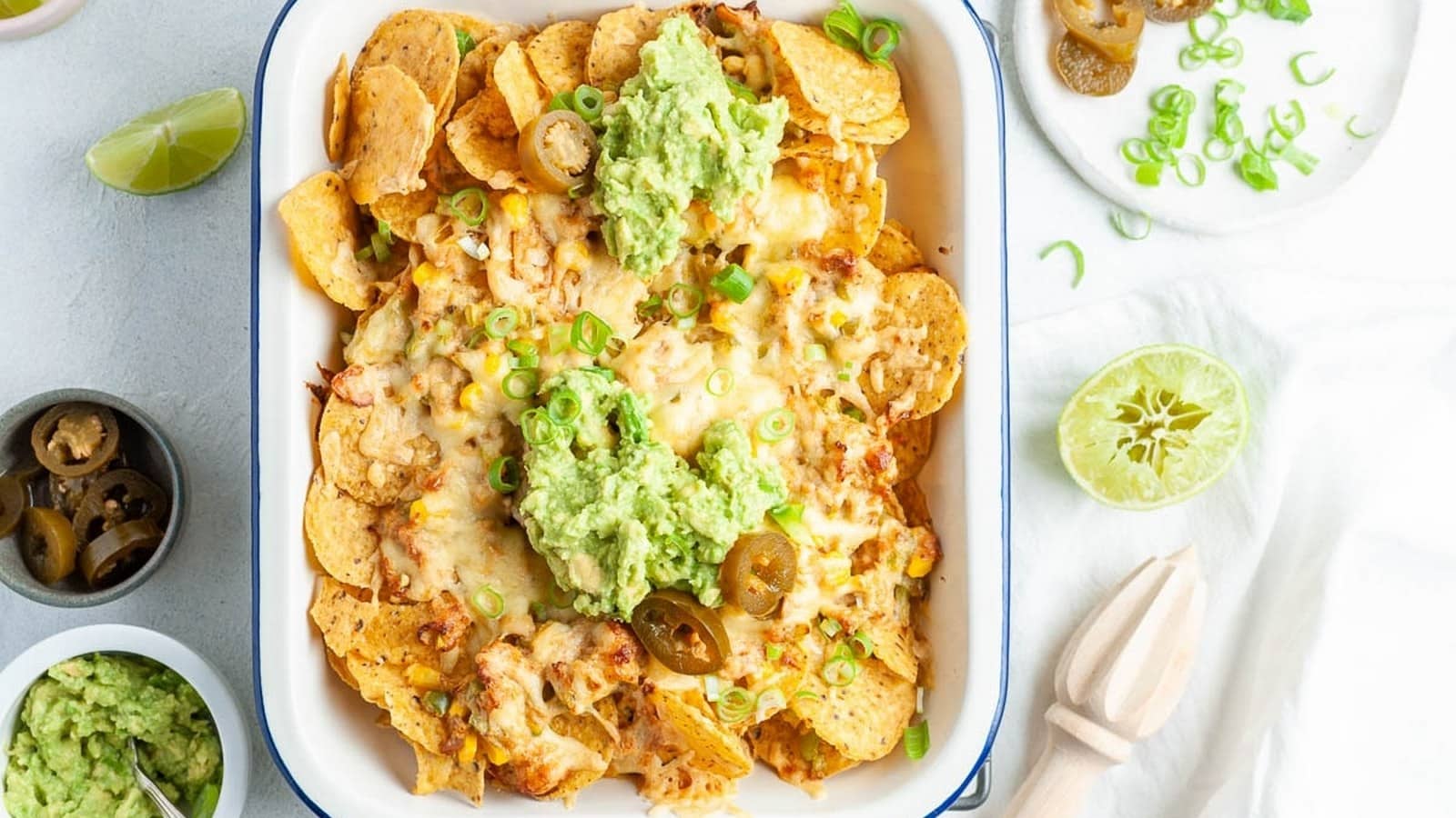 A dish of baked nachos topped with melted cheese, guacamole, jalapeños, and green onions, on a light surface with lime halves and a bowl of guacamole nearby.