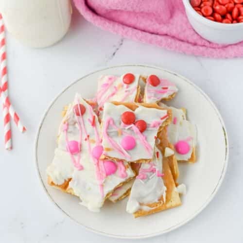 A plate of dessert squares topped with white chocolate, pink drizzle, and red candy pieces. A pink cloth and bowl of red candies are in the background.