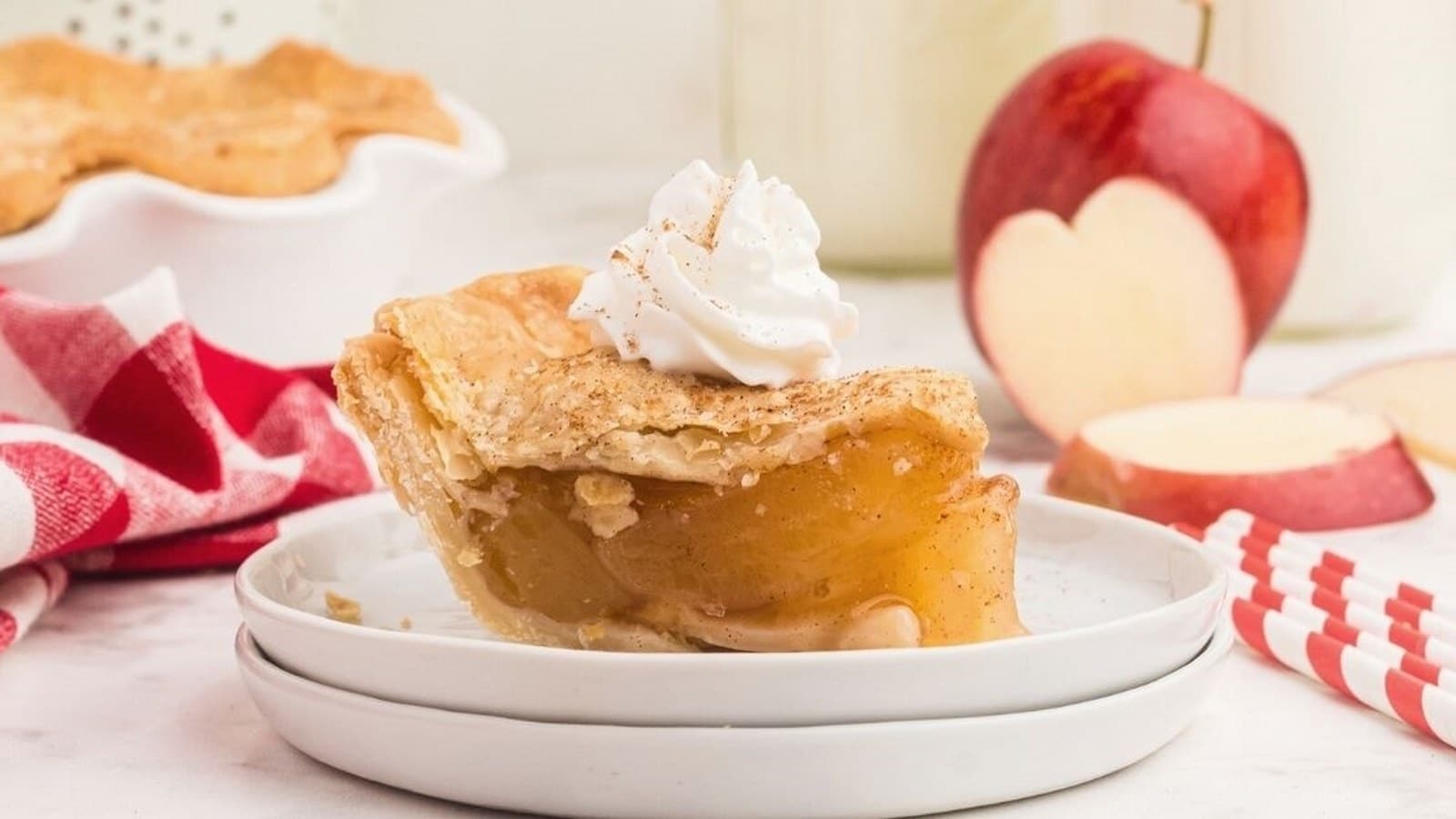 Slice of apple pie with whipped cream on top, placed on a stack of plates. A red apple, apple slices, and a red-checkered cloth are in the background.