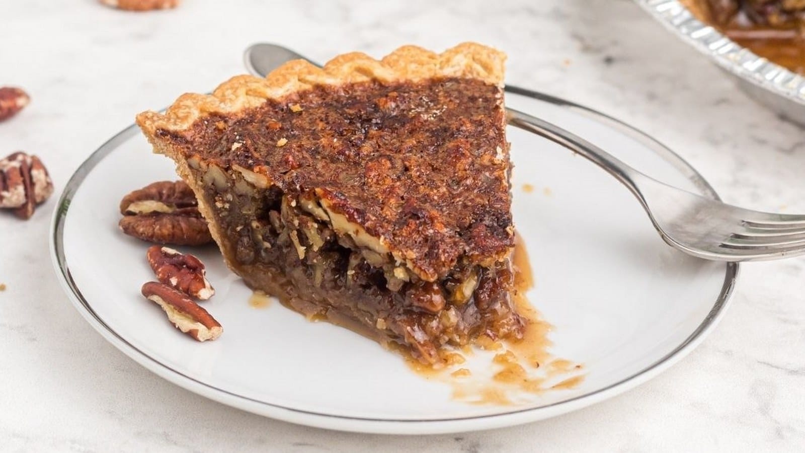 Slice of pecan pie on a white plate with a fork beside it; pecans are scattered on the marble surface.