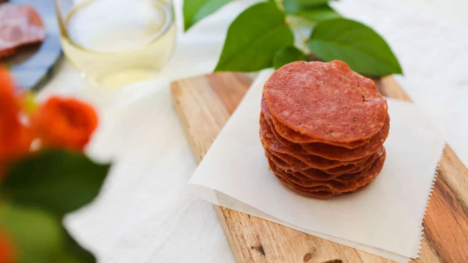 A stack of thinly sliced cured meat on parchment paper, placed on a wooden board, with a glass and green leaves in the background.