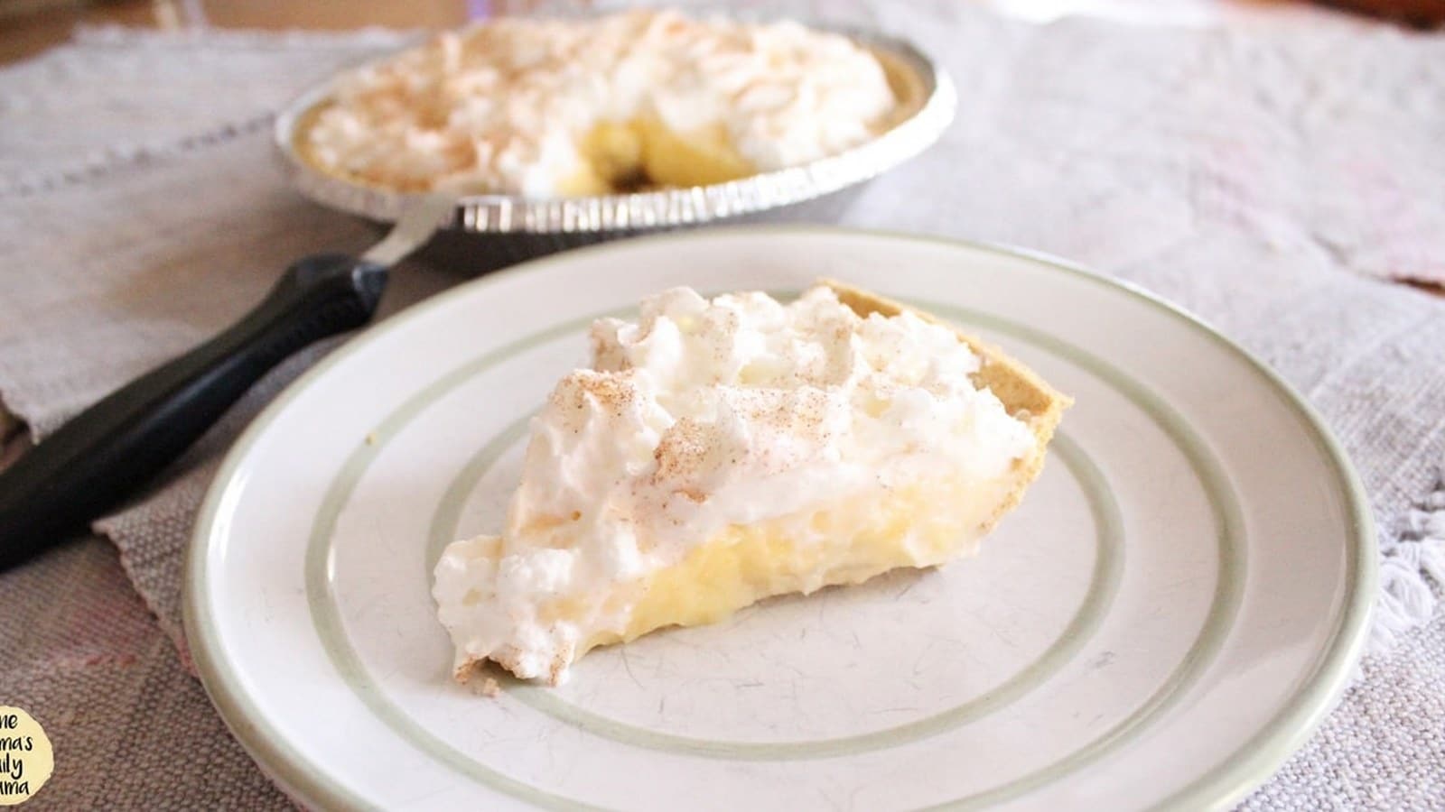 A slice of pie with a creamy filling and fluffy topping on a white plate, with the rest of the pie in a tin in the background.