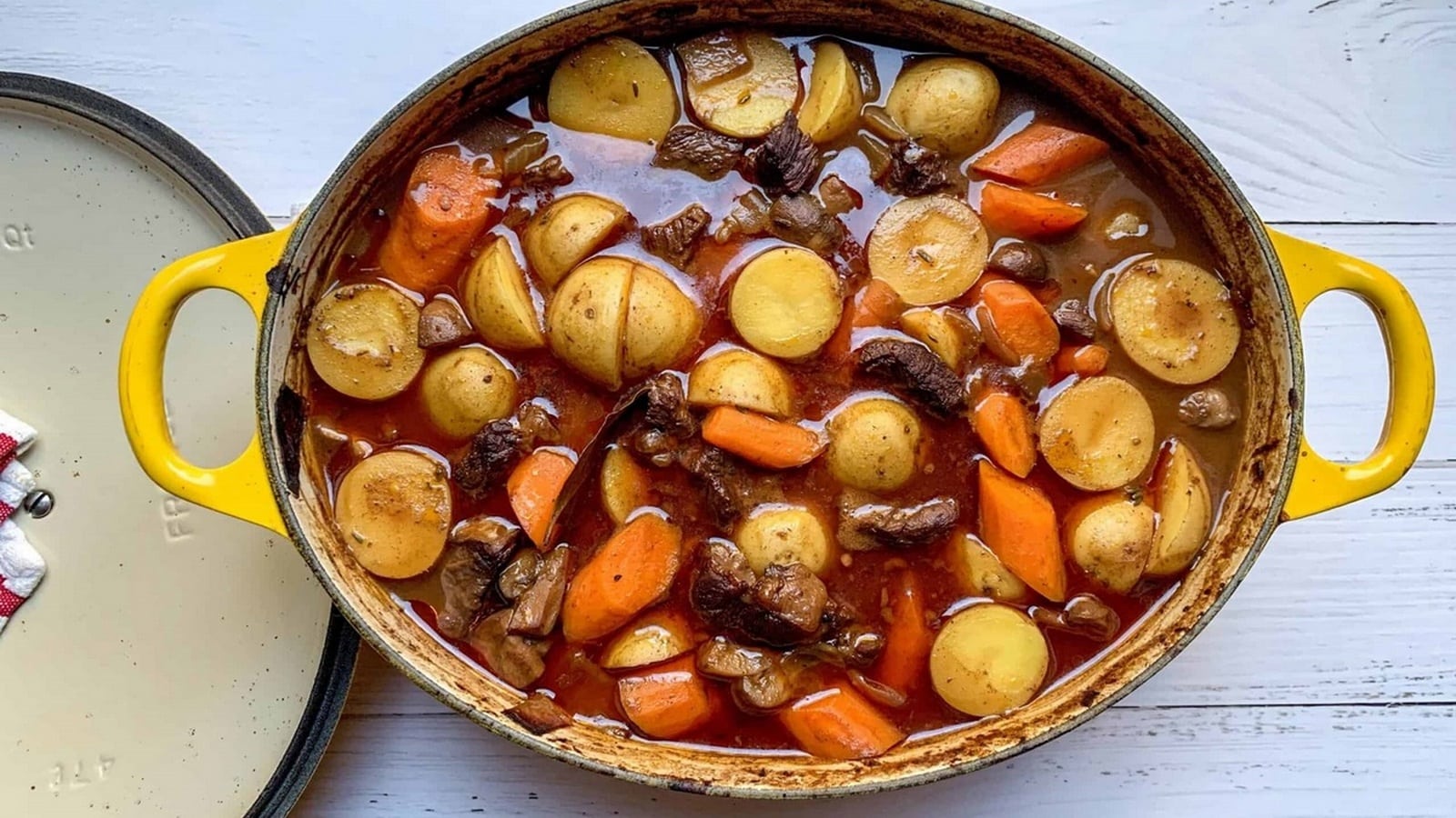 A yellow pot filled with beef stew, containing pieces of beef, potatoes, and carrots in a brown sauce on a wooden surface.