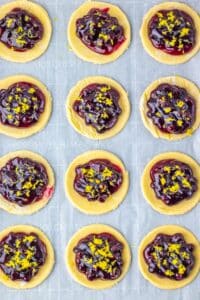 Twelve cookies with a golden base topped with glossy berry compote and sprinkled with lemon zest, arranged in a grid pattern on parchment paper.