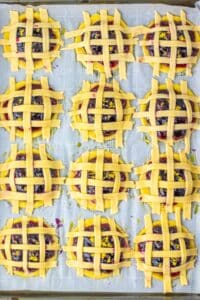 Twelve mini lattice pies with golden crust and blueberry filling on a parchment-lined baking sheet.