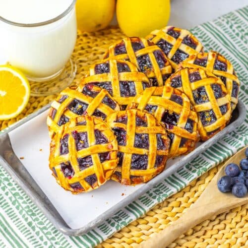 A tray of small round blueberry pies with a lattice crust on a green and white cloth, surrounded by lemons, a glass of milk, and blueberries on a wooden spoon.