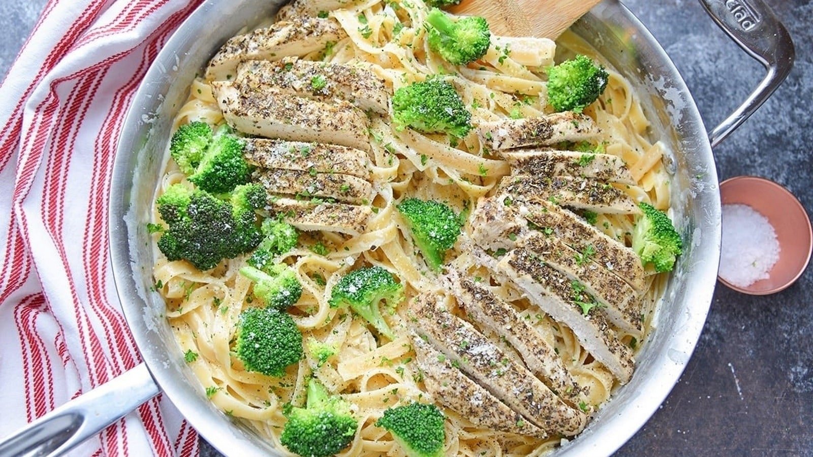 A skillet filled with fettuccine pasta, sliced seasoned chicken, and broccoli, with a side of salt in a small dish and a striped cloth beside the pan.