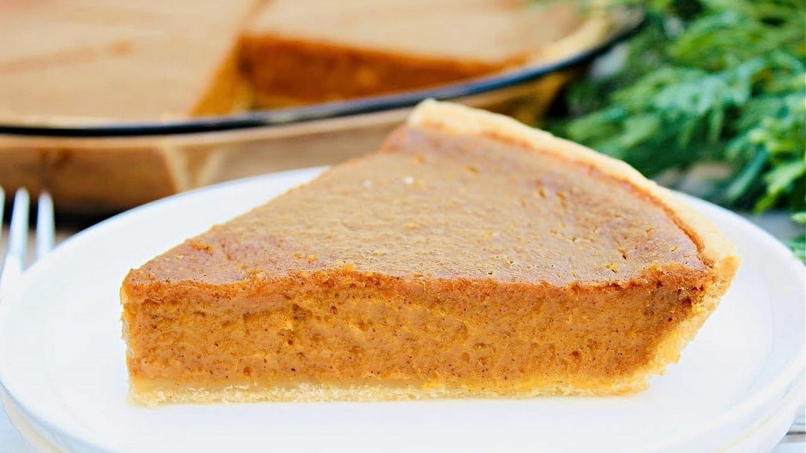 A slice of pumpkin pie on a white plate, with a fork beside it and greenery in the background.