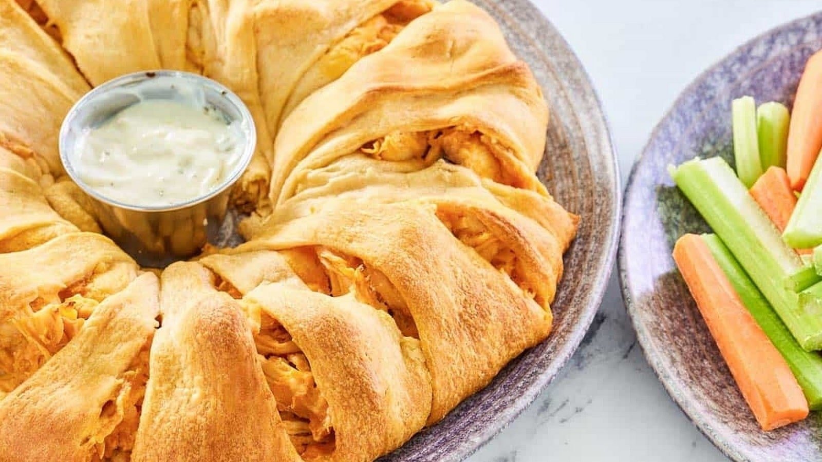 A crescent roll buffalo chicken ring with a cup of dipping sauce in the center, served on a plate. Nearby, a plate with celery and carrot sticks is visible.