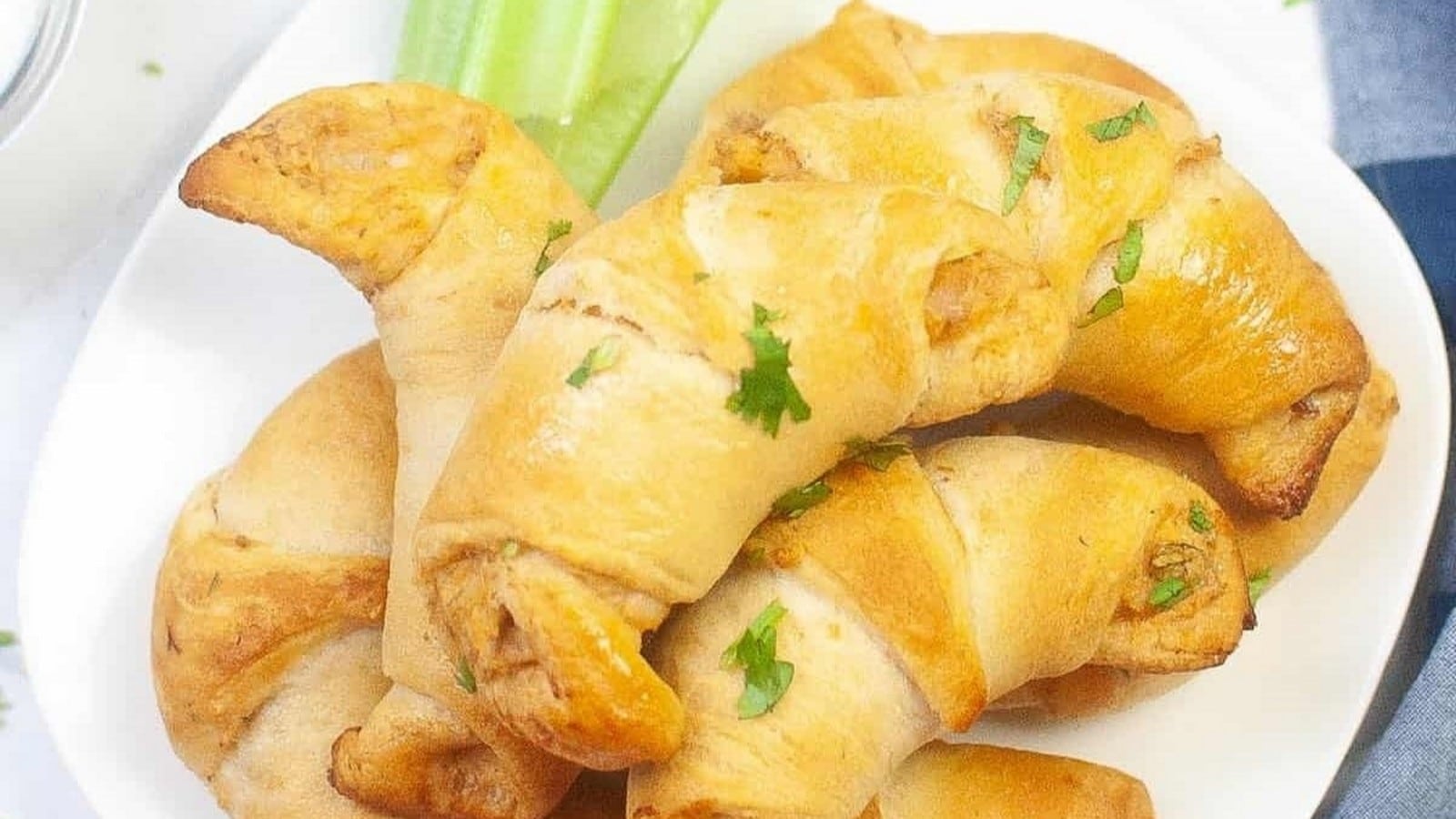 A plate of golden-brown crescent rolls garnished with parsley, accompanied by a celery stick.