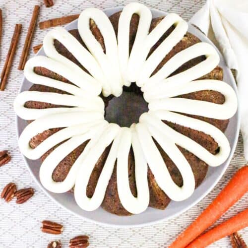 A carrot bundt cake with white icing drizzle is on a plate. Cinnamon sticks, carrots, and pecans are around the plate.