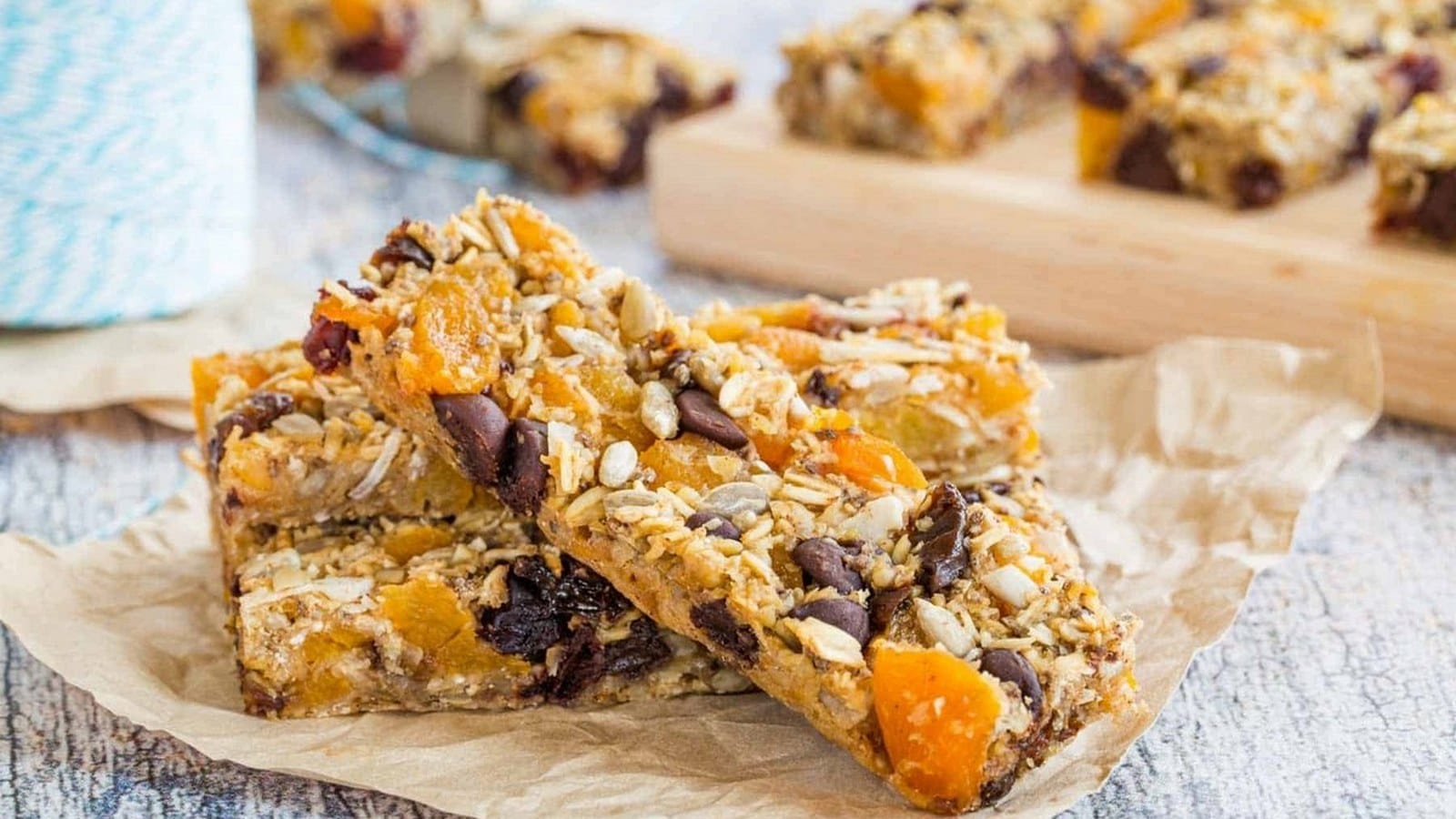 Homemade granola bars with dried fruits, nuts, and chocolate chunks on parchment paper, with more bars in the background on a wooden board.