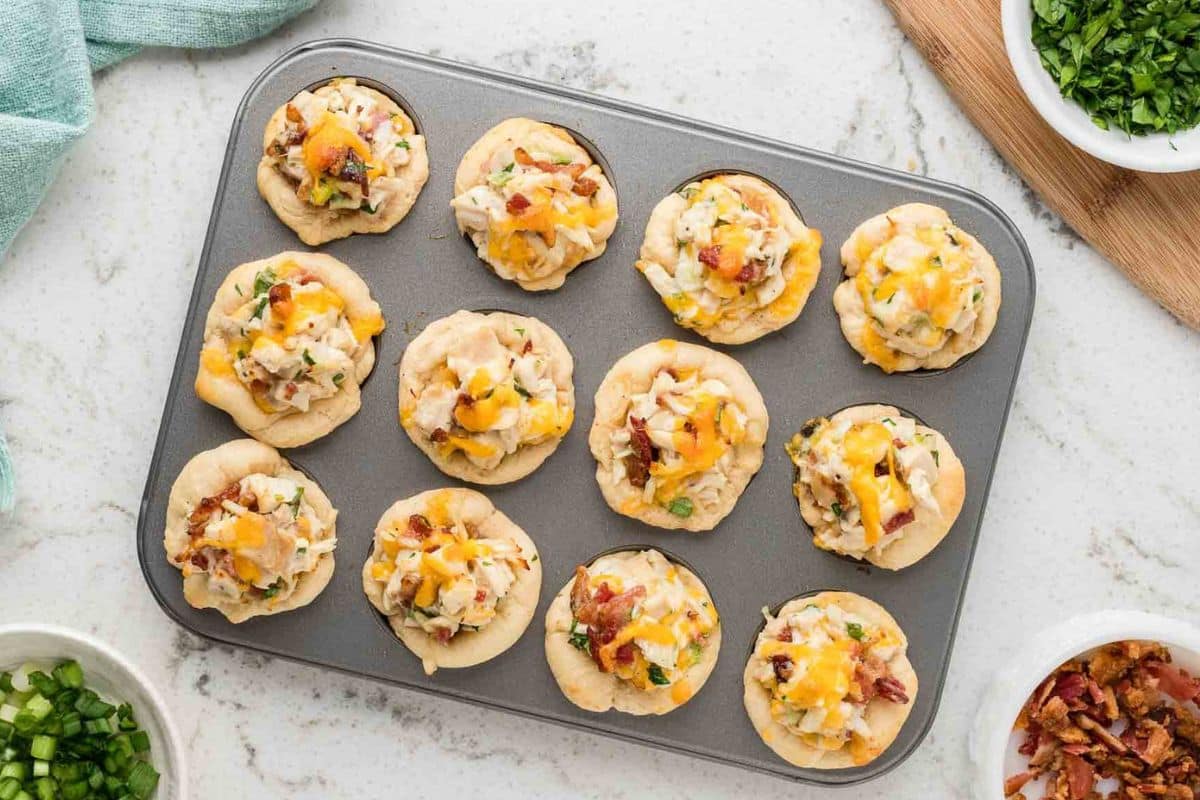 Twelve mini chicken biscuit cups with cheese and herbs in a muffin tin on a marble surface, surrounded by fresh ingredients in bowls.