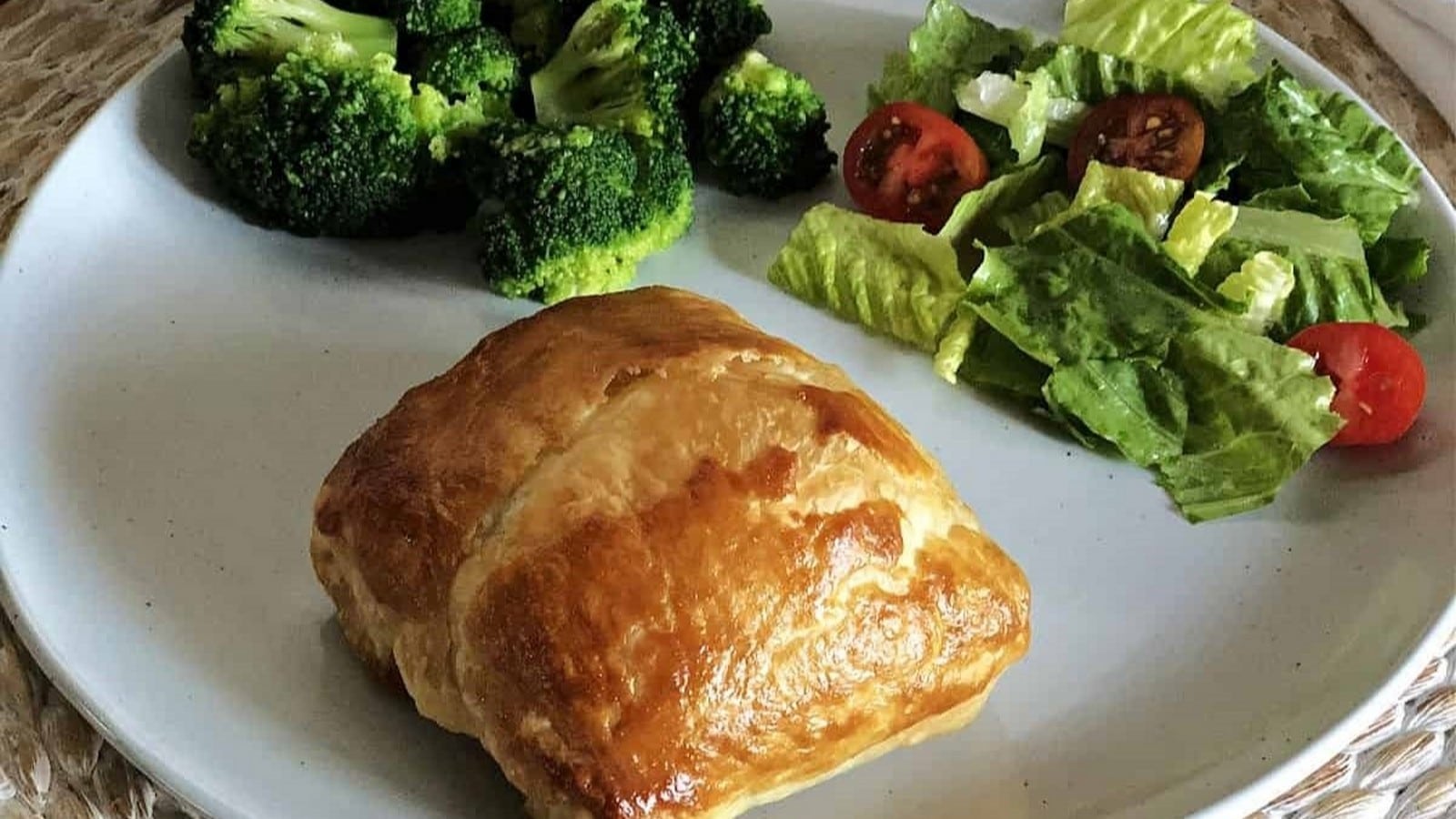 A puff pastry parcel on a plate with broccoli and a green salad with tomatoes.