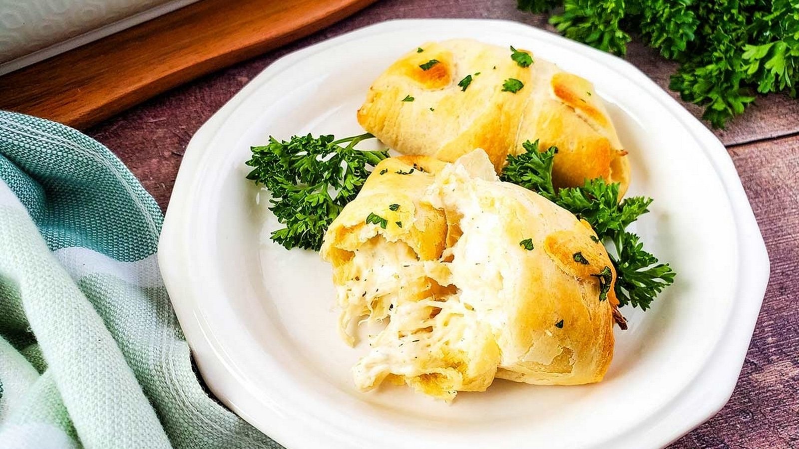 Two cheesy stuffed pastries on a white plate, garnished with fresh parsley, with a wooden board and green cloth nearby.