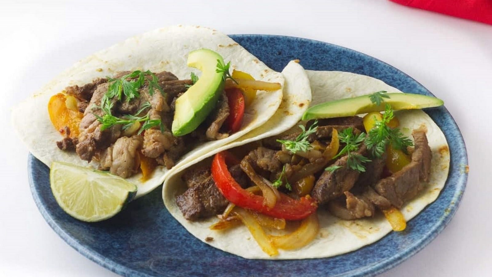 Two soft tacos with grilled beef, avocado slices, and bell peppers on a blue plate, garnished with cilantro, with a lime wedge nearby.