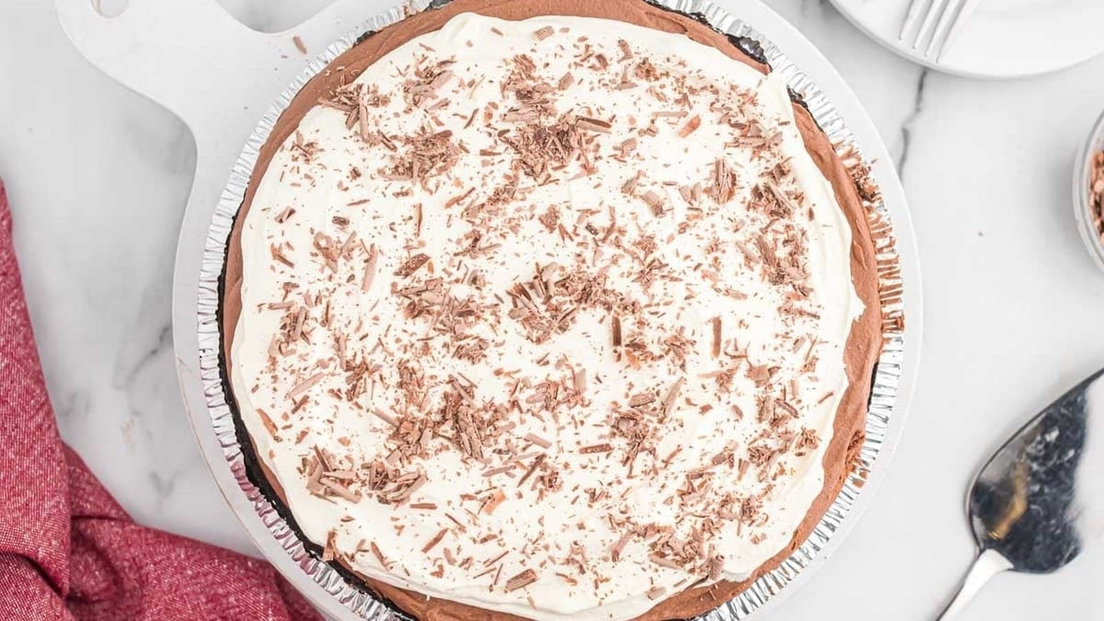 Cream pie topped with chocolate shavings on a white surface, next to a red cloth and a serving spatula.