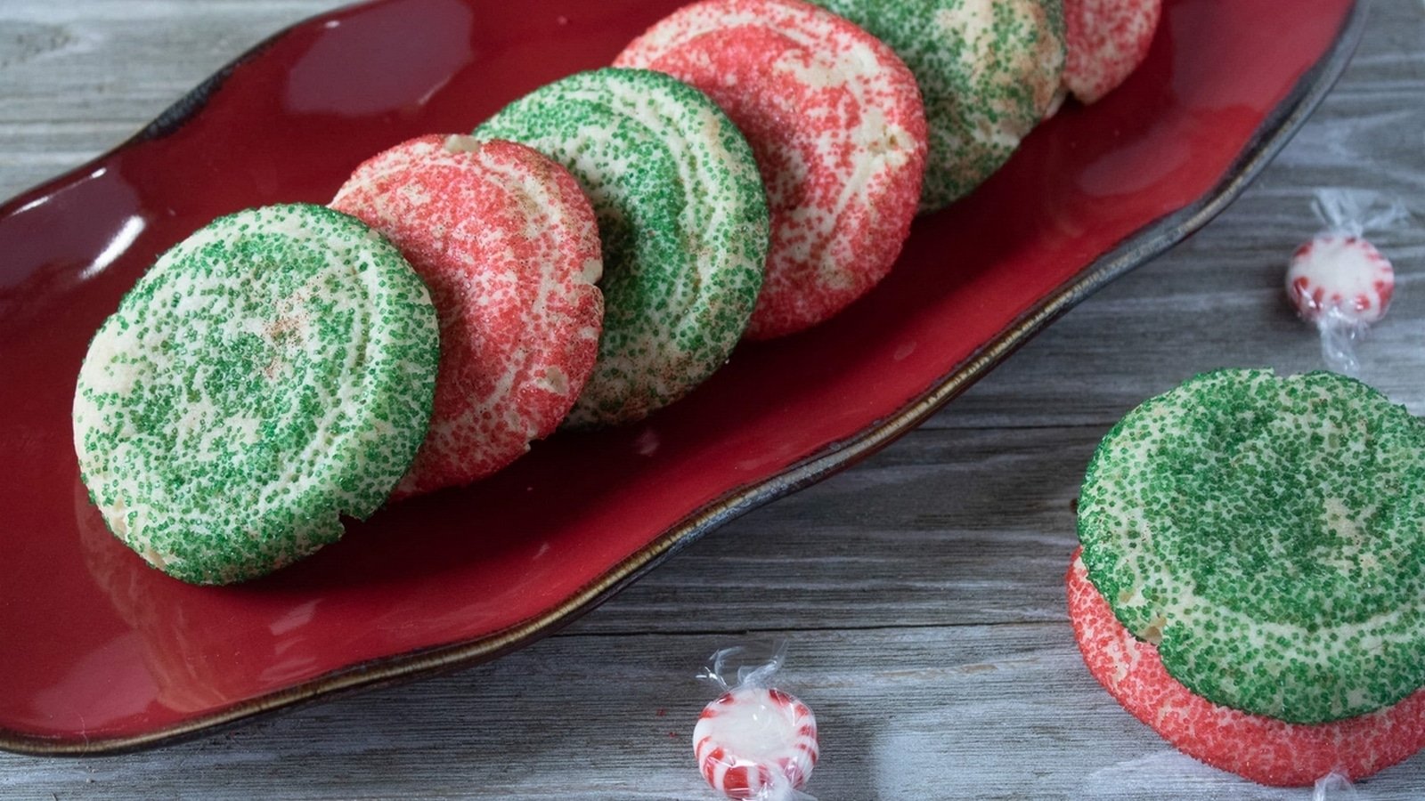 Red and green sugar cookies arranged on a red platter, with two peppermint candies nearby on a wooden surface.