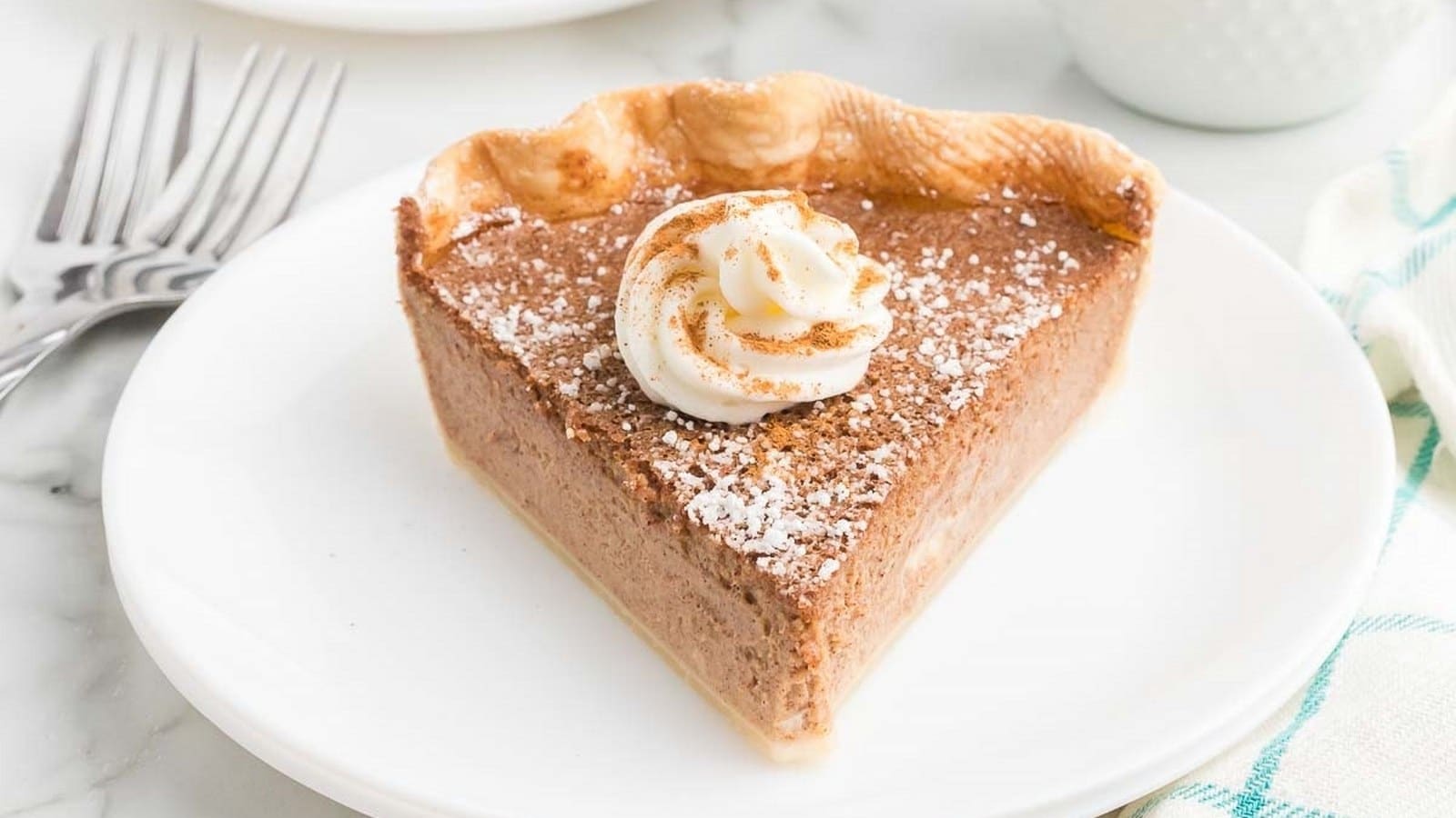 A slice of pie on a white plate, topped with whipped cream and a sprinkle of powdered sugar, with a crimped crust edge. Forks are visible in the background.