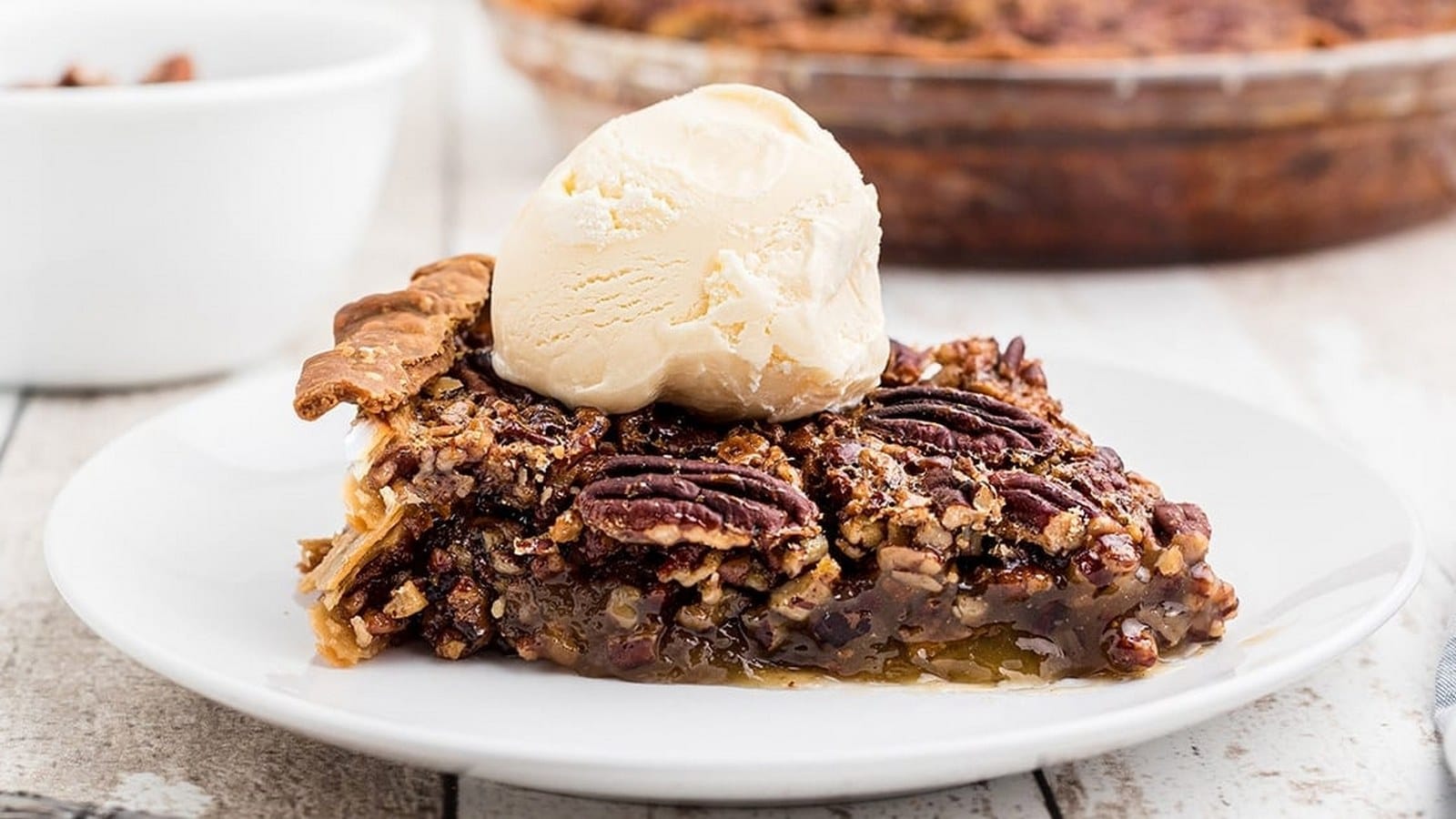 Slice of pecan pie topped with a scoop of vanilla ice cream on a white plate.