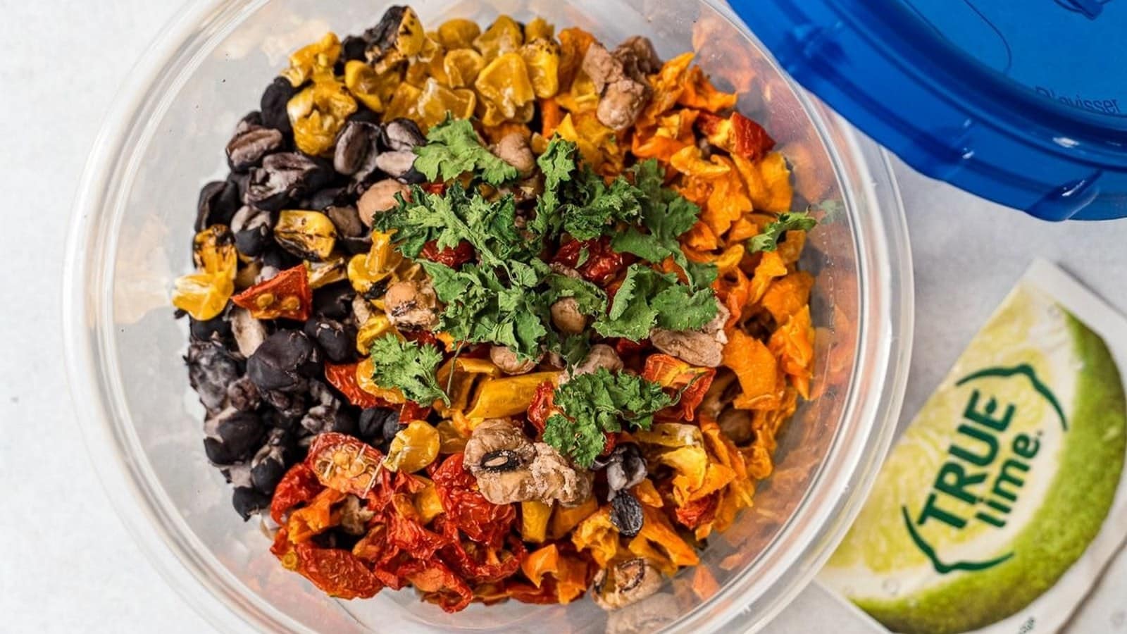 A plastic bowl filled with assorted dried vegetables and herbs next to a pack of True Lime on a white surface.
