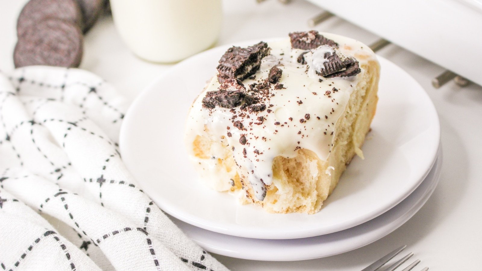 A cinnamon roll topped with white icing and crushed chocolate cookies on a white plate, next to a napkin, a fork, and milk.