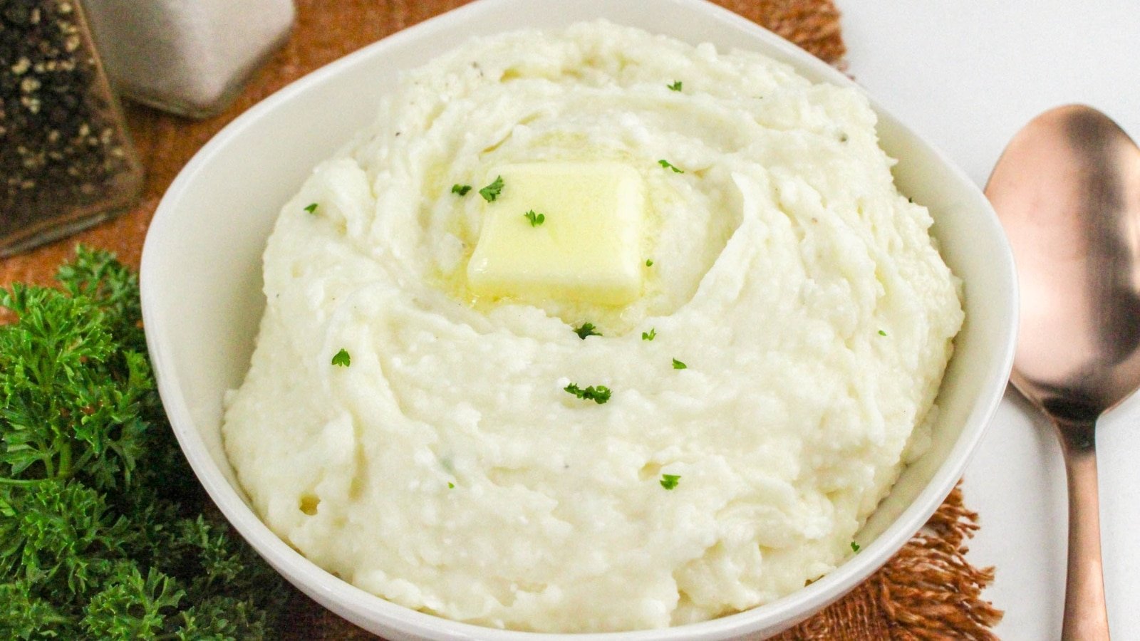 Bowl of mashed potatoes with a pat of butter on top, garnished with parsley. A spoon and a sprig of parsley are next to the bowl.