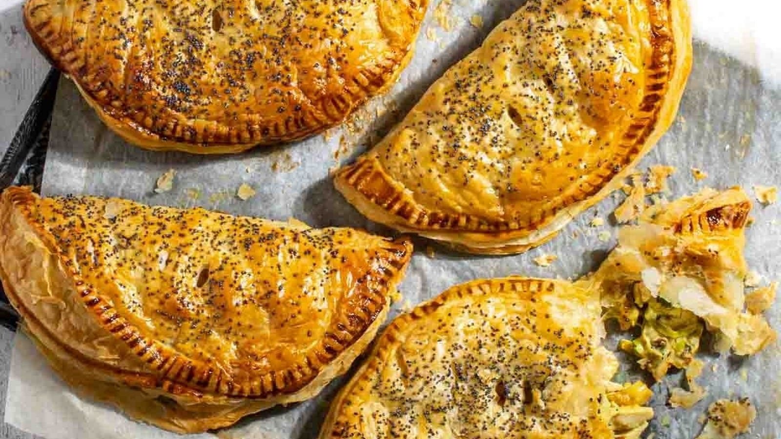 Four baked pastries with golden, flaky crusts and poppy seeds are arranged on parchment paper. One pastry is cut open, revealing a green vegetable filling.