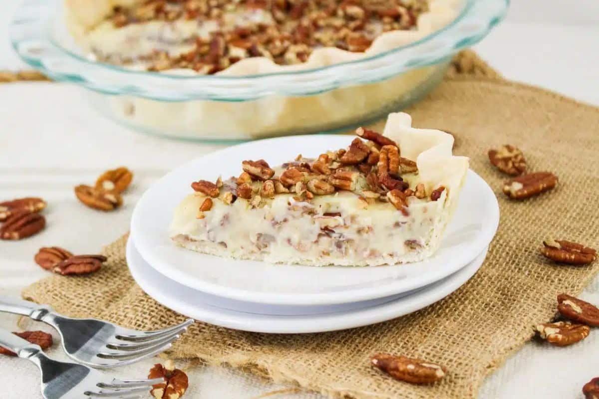 A slice of pecan pie on white plates, garnished with chopped pecans. The pie is placed on a burlap cloth with whole pecans scattered around. Two forks are beside it.