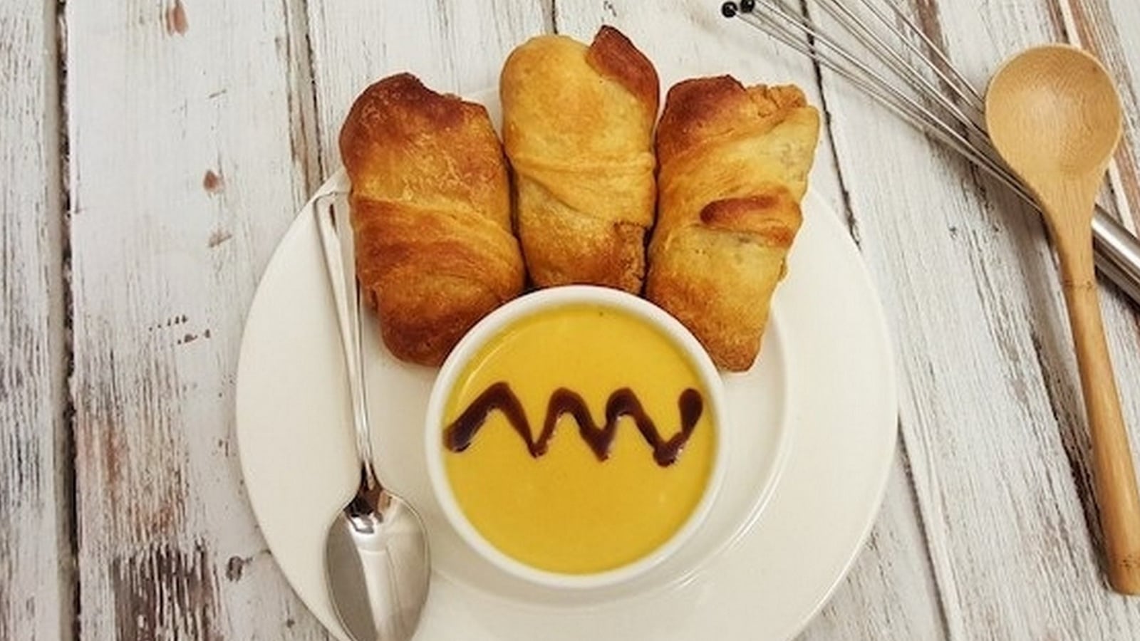 A plate with three crescent rolls and a bowl of soup with sauce on top. A metal whisk, wooden spoons, and a spoon are beside the plate on a wooden surface.