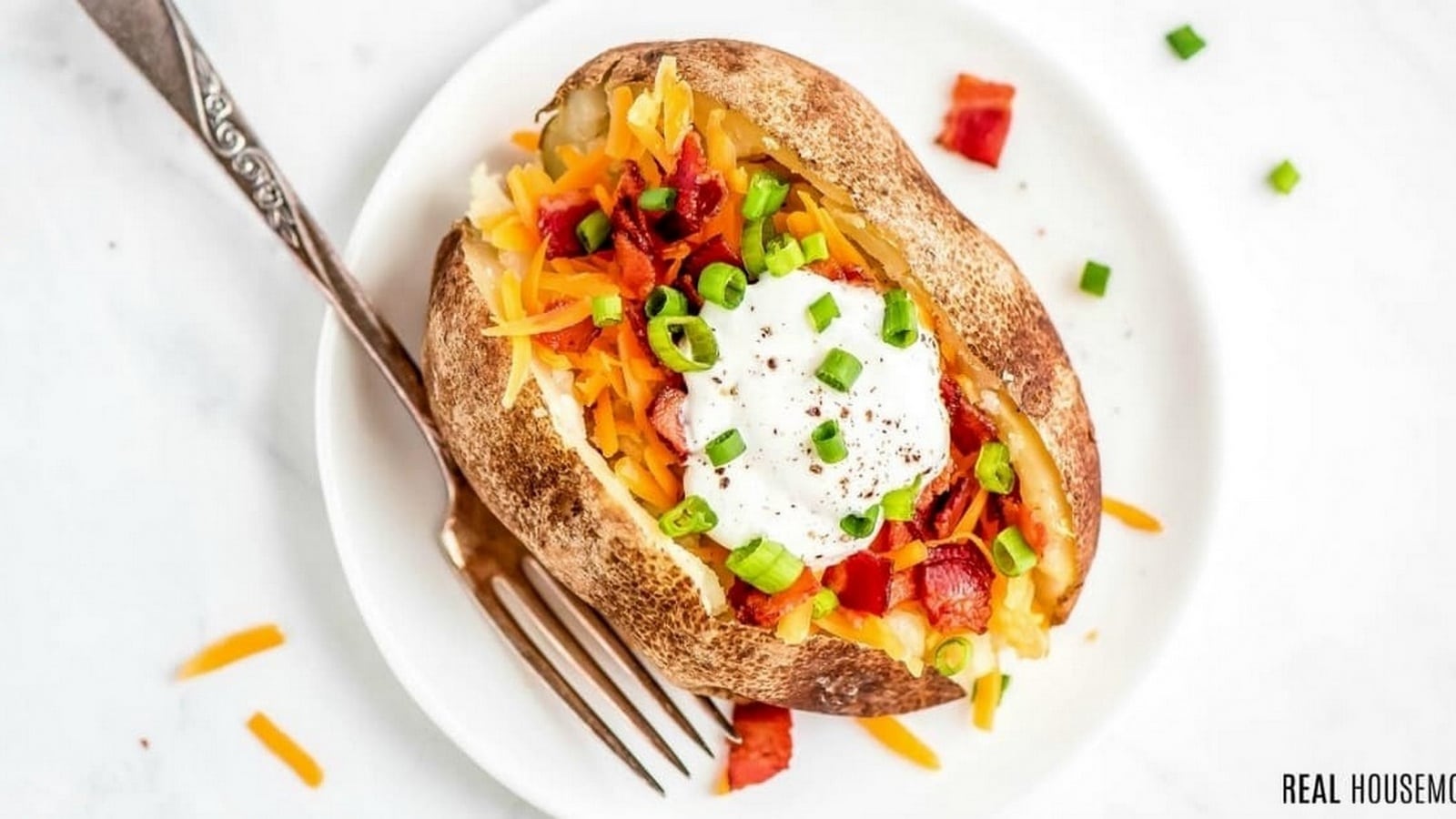 Baked potato topped with sour cream, shredded cheese, chopped green onions, and bacon bits on a white plate with a fork.