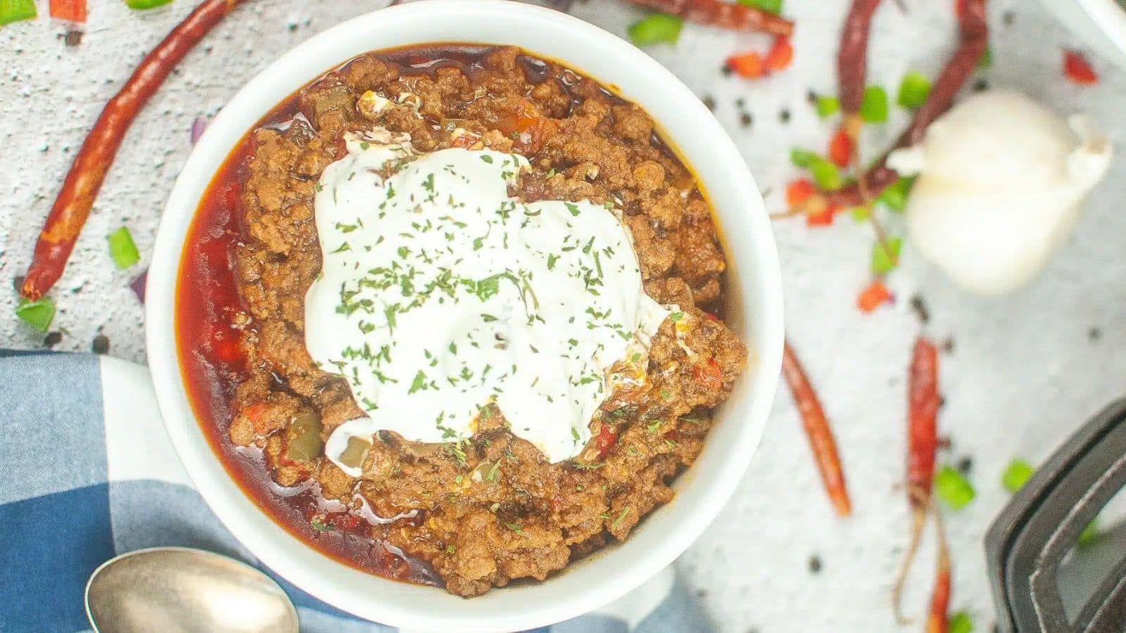 A bowl of chili with ground meat, topped with sour cream and herbs. Surrounding items include whole garlic, red chili peppers, and chopped green onions. A spoon and a checkered cloth are nearby.