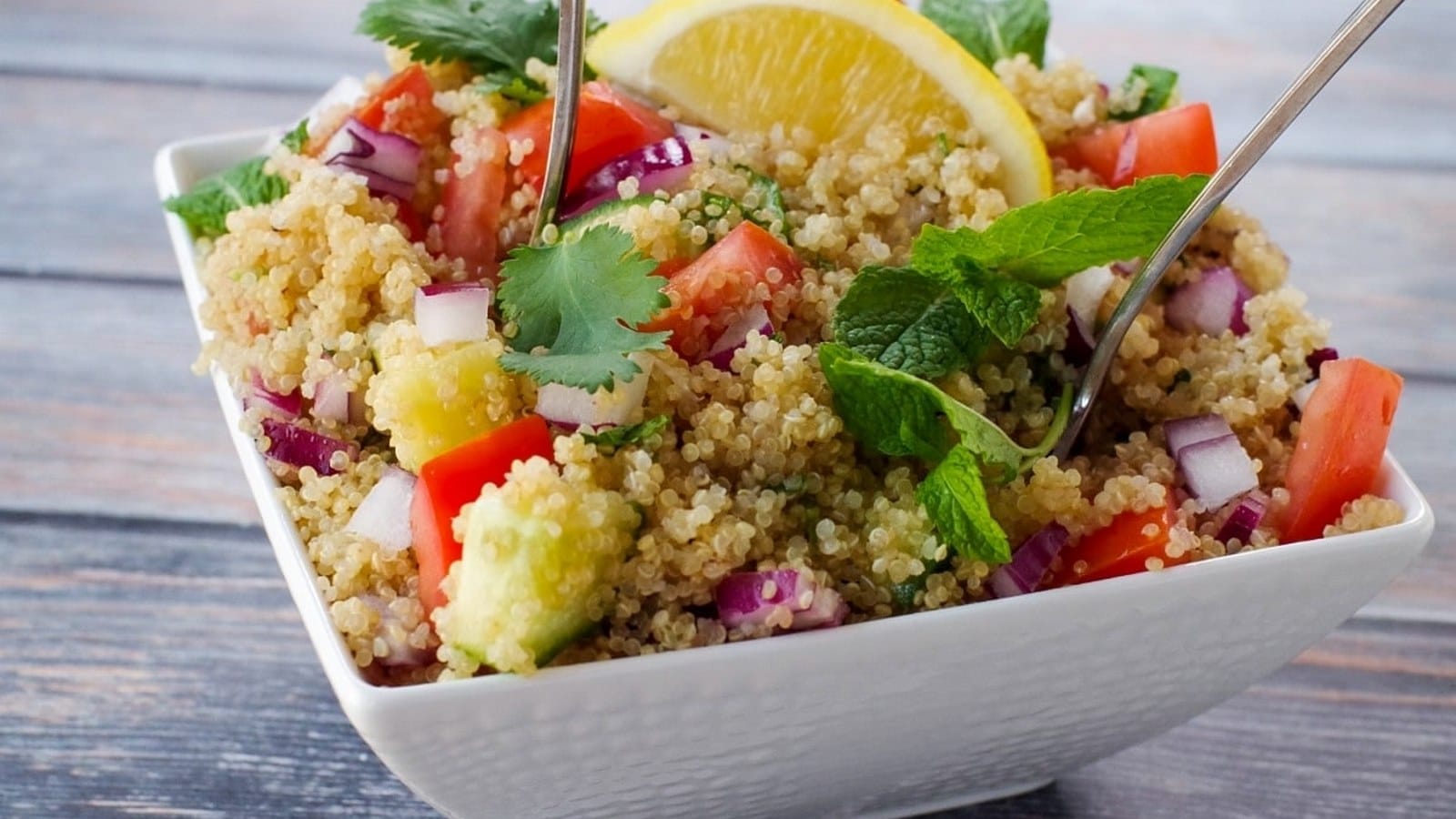 A bowl of quinoa salad with diced tomatoes, red onions, cucumber, cilantro, mint, and a lemon wedge, served with two forks.