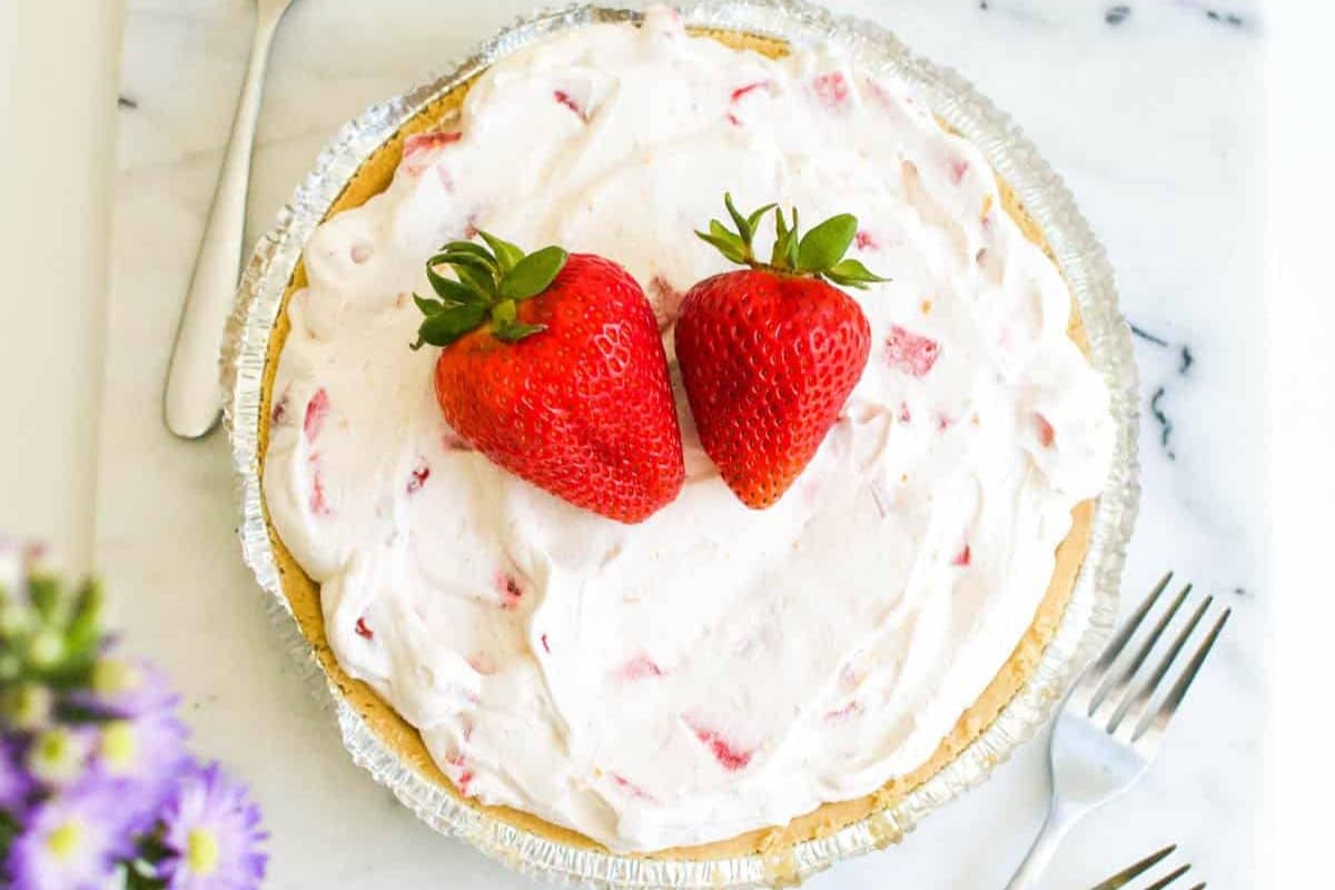 A pie topped with whipped cream and two strawberries in a metal pie tin. Two forks are on the side, and purple flowers are visible in the bottom left corner.