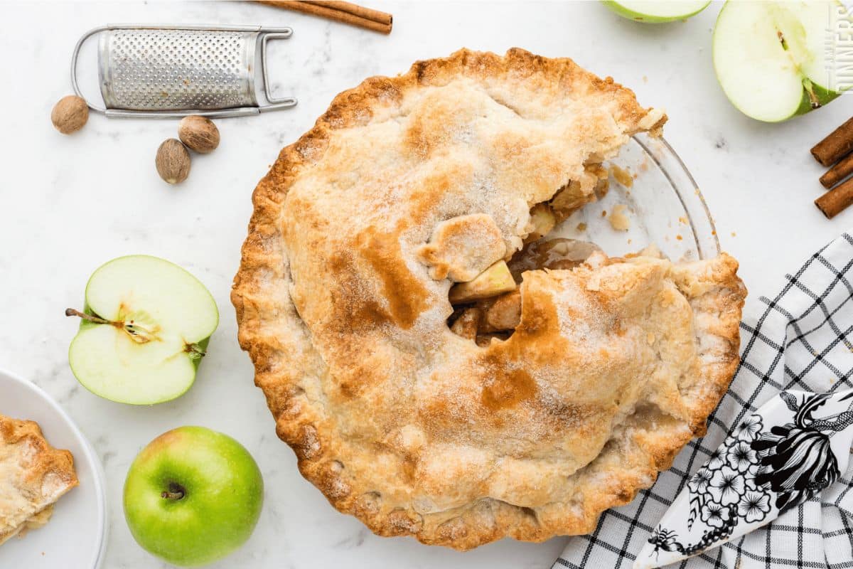 Apple pie with a slice removed on a marble surface, surrounded by whole and sliced green apples, cinnamon sticks, a grater, and a patterned cloth.