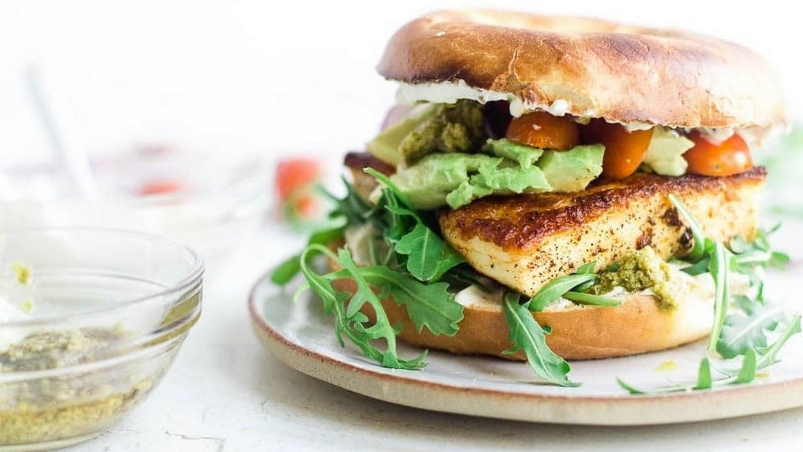 Bagel sandwich with grilled chicken, avocado, cherry tomatoes, arugula, and pesto on a plate. Small glass bowl with dressing nearby.