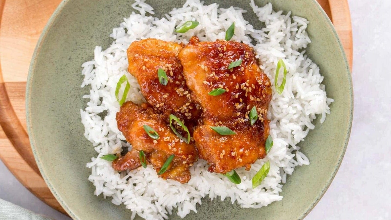 A dish of crispy chicken pieces topped with sesame seeds and green onions, served over a bed of white rice in a green bowl.