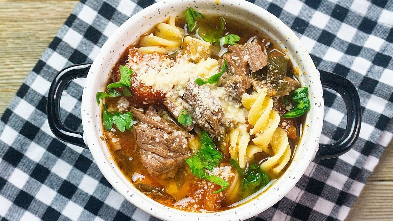 A bowl of soup with beef chunks, pasta, vegetables, and grated cheese, garnished with parsley, placed on a black and white checkered cloth.