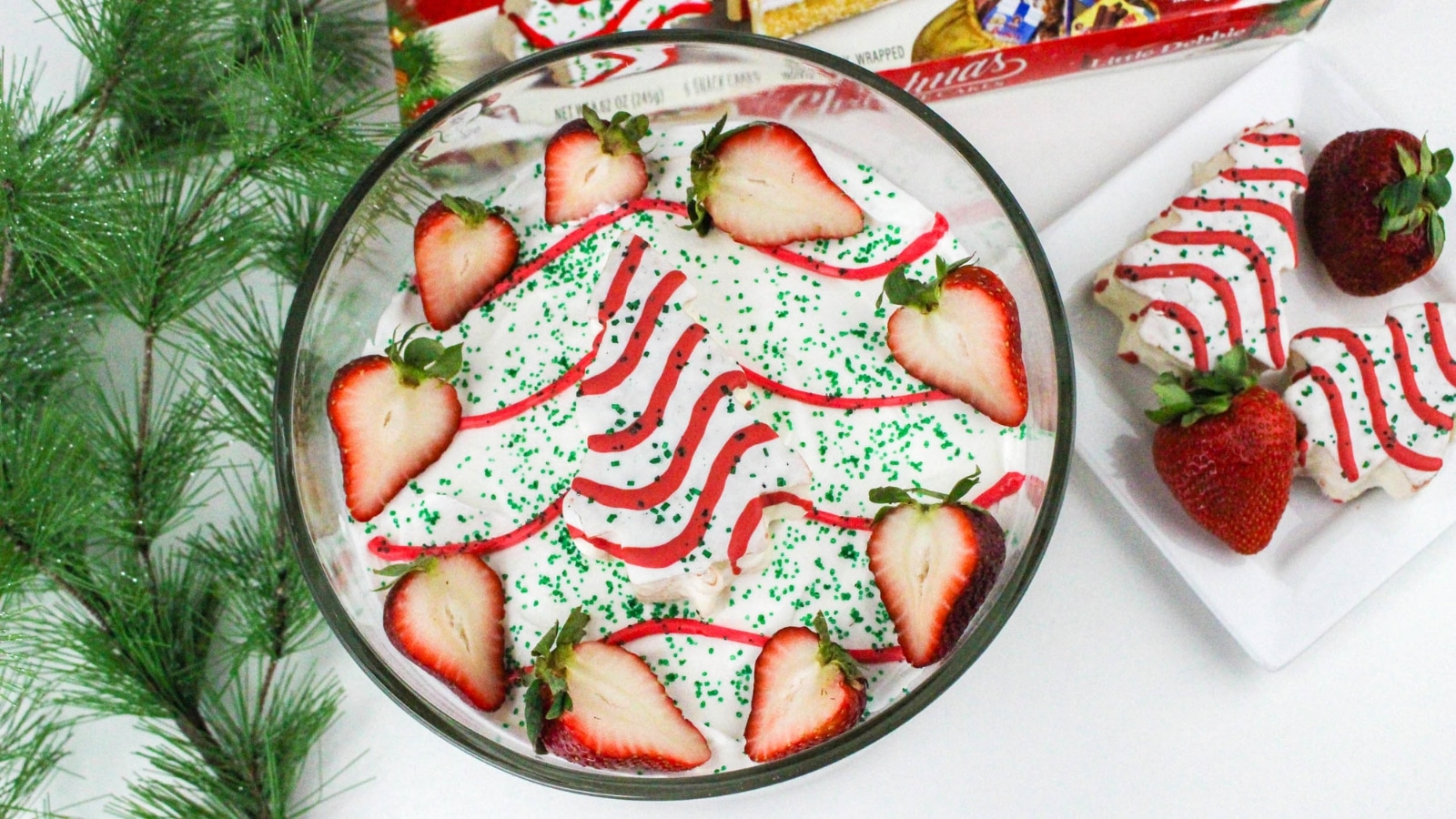 A bowl of dessert topped with sliced strawberries, green sprinkles, and red icing, next to a plate with similar decorated treats. Pine branches are on the side.