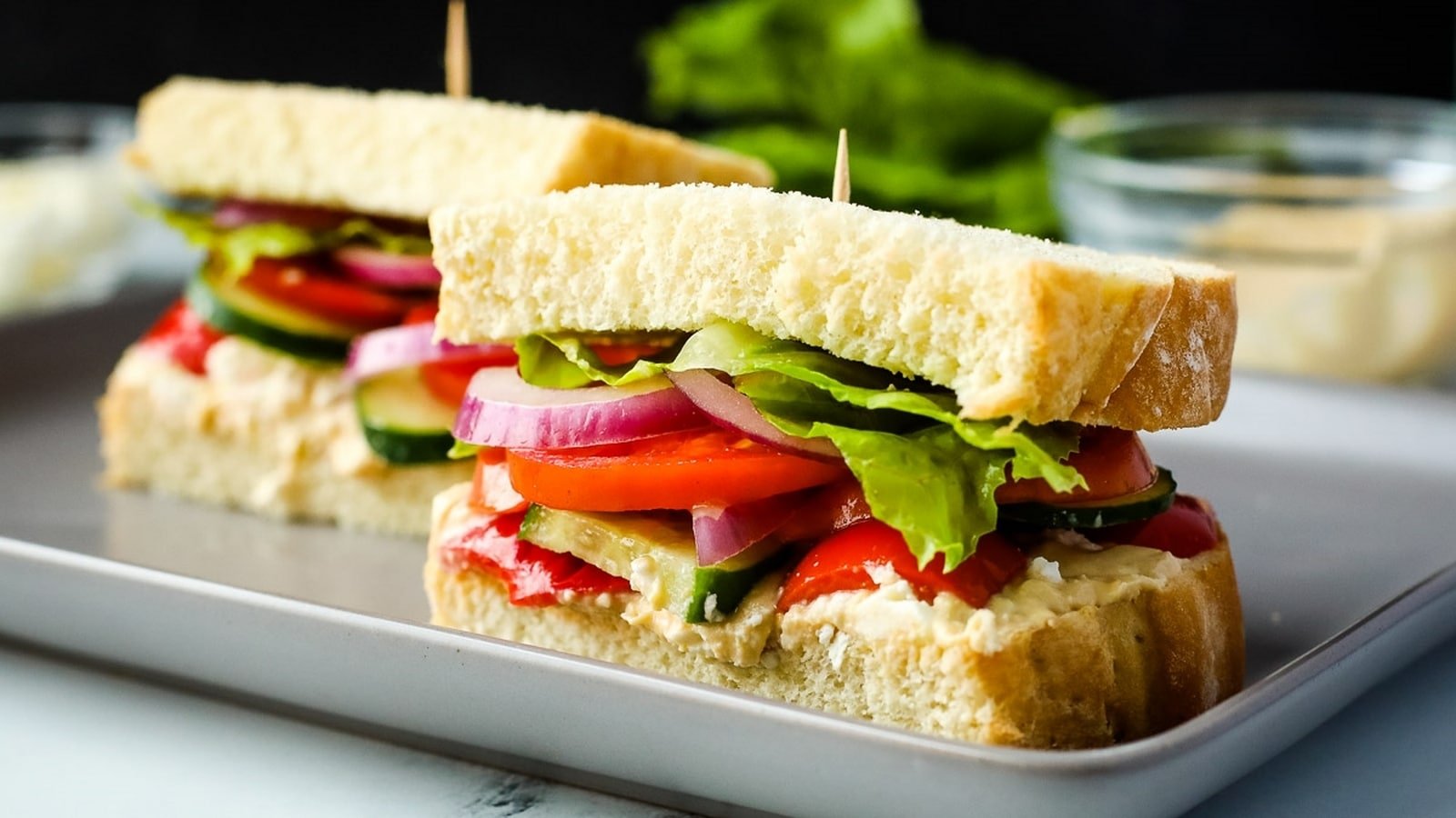 Two sandwiches on a plate, filled with lettuce, tomato, cucumber, red onion, and a spread, secured with toothpicks.