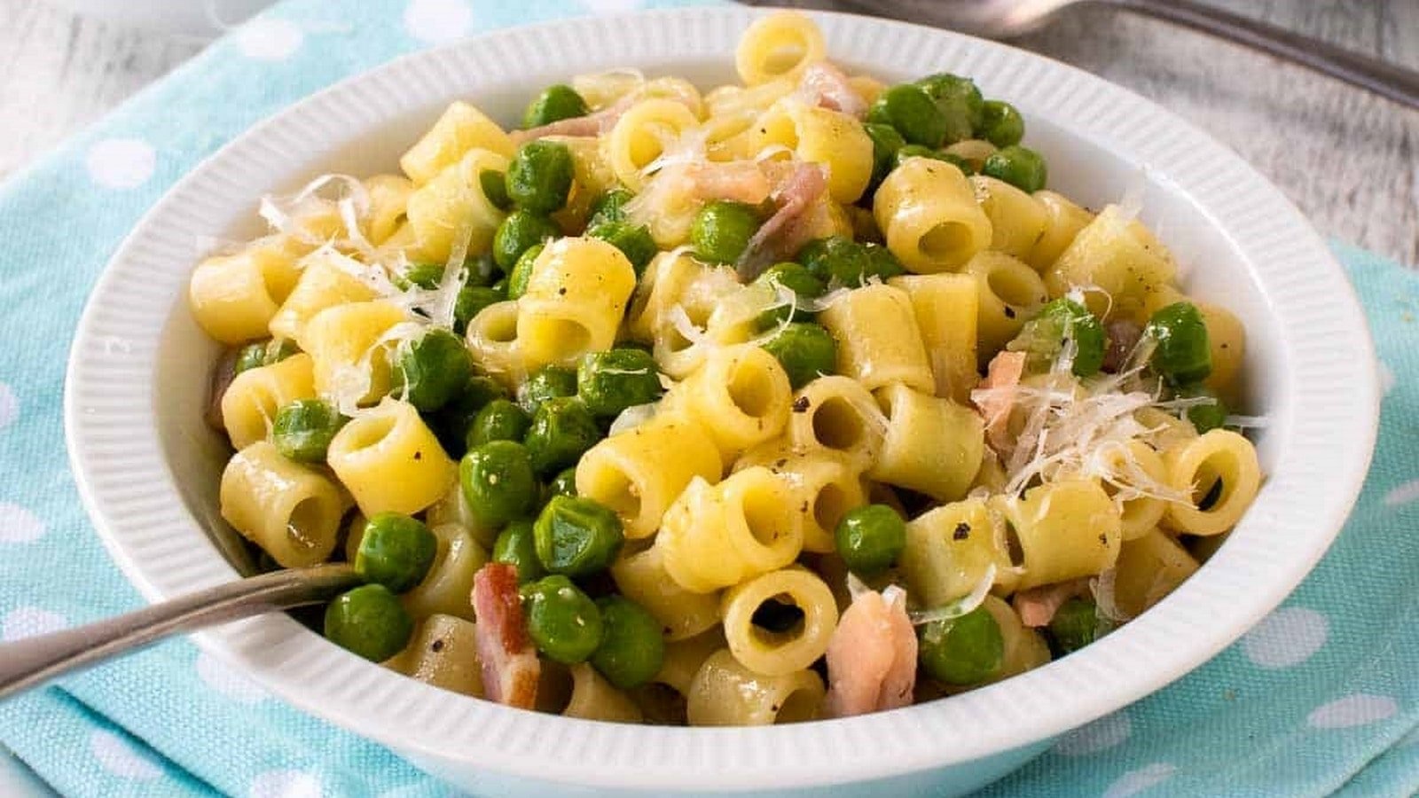 A bowl of pasta with peas, grated cheese, and bacon pieces, served with a spoon on a blue polka dot cloth.