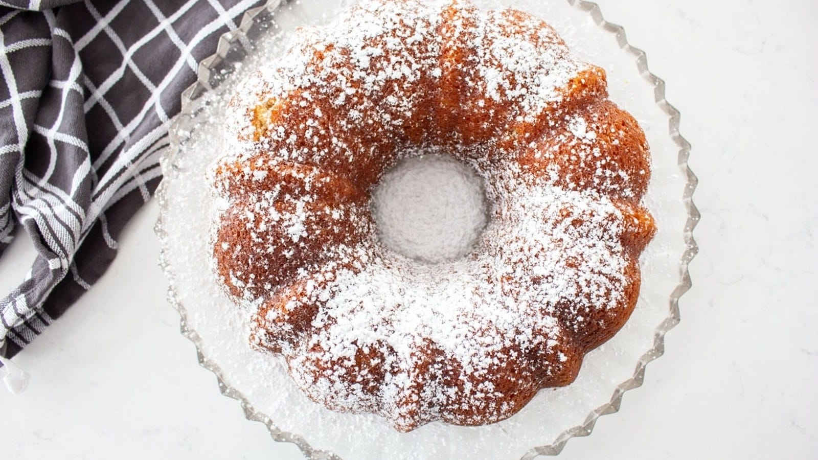 A Bundt cake dusted with powdered sugar on a glass cake stand, next to a black and white checkered cloth.