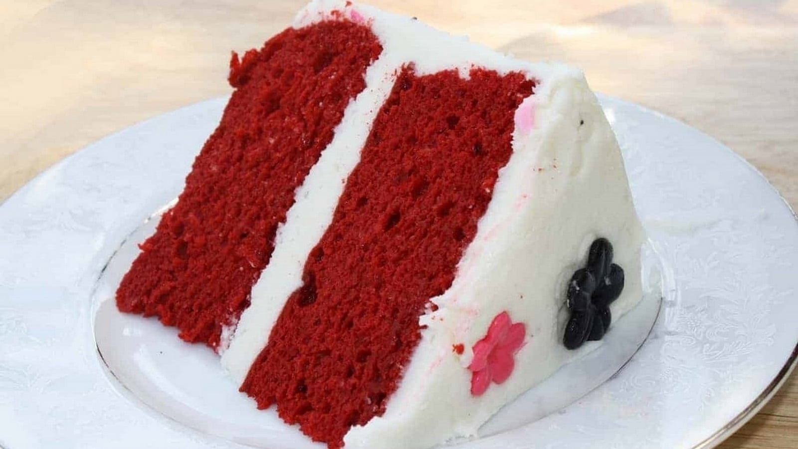 A slice of red velvet cake with white frosting on a white plate. Decorative pink and black flower shapes are on the side.