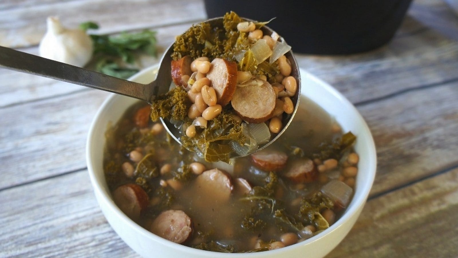 A spoonful of soup with slices of sausage, beans, greens, and broth being served into a white bowl on a wooden table. Garlic and herbs are in the background.