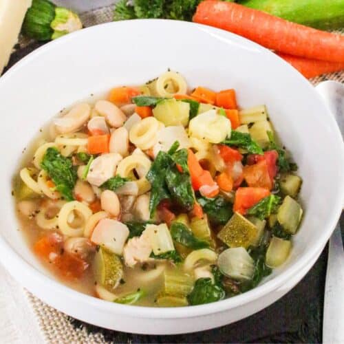 A bowl of vegetable soup with loop-shaped pasta, white beans, carrots, celery, spinach, and diced tomatoes. Fresh vegetables are visible in the background.