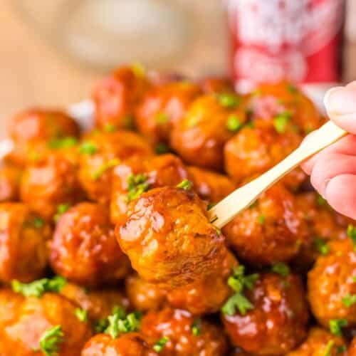 Close-up of a hand holding a small skewer with a glazed meatball garnished with herbs, above a plate of similar meatballs.