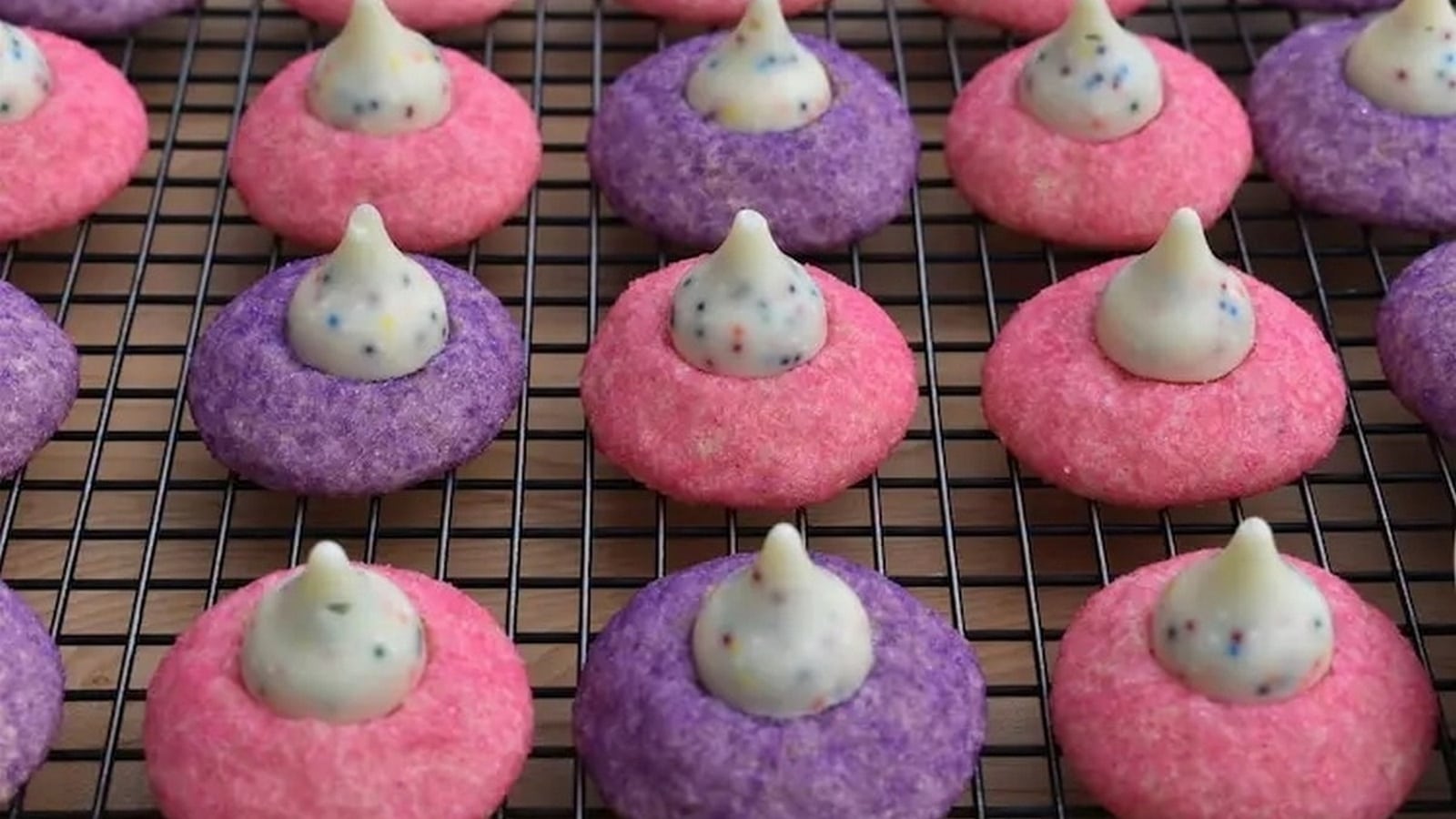 Pink and purple cookies with white chocolate and sprinkles on top arranged in rows on a cooling rack.