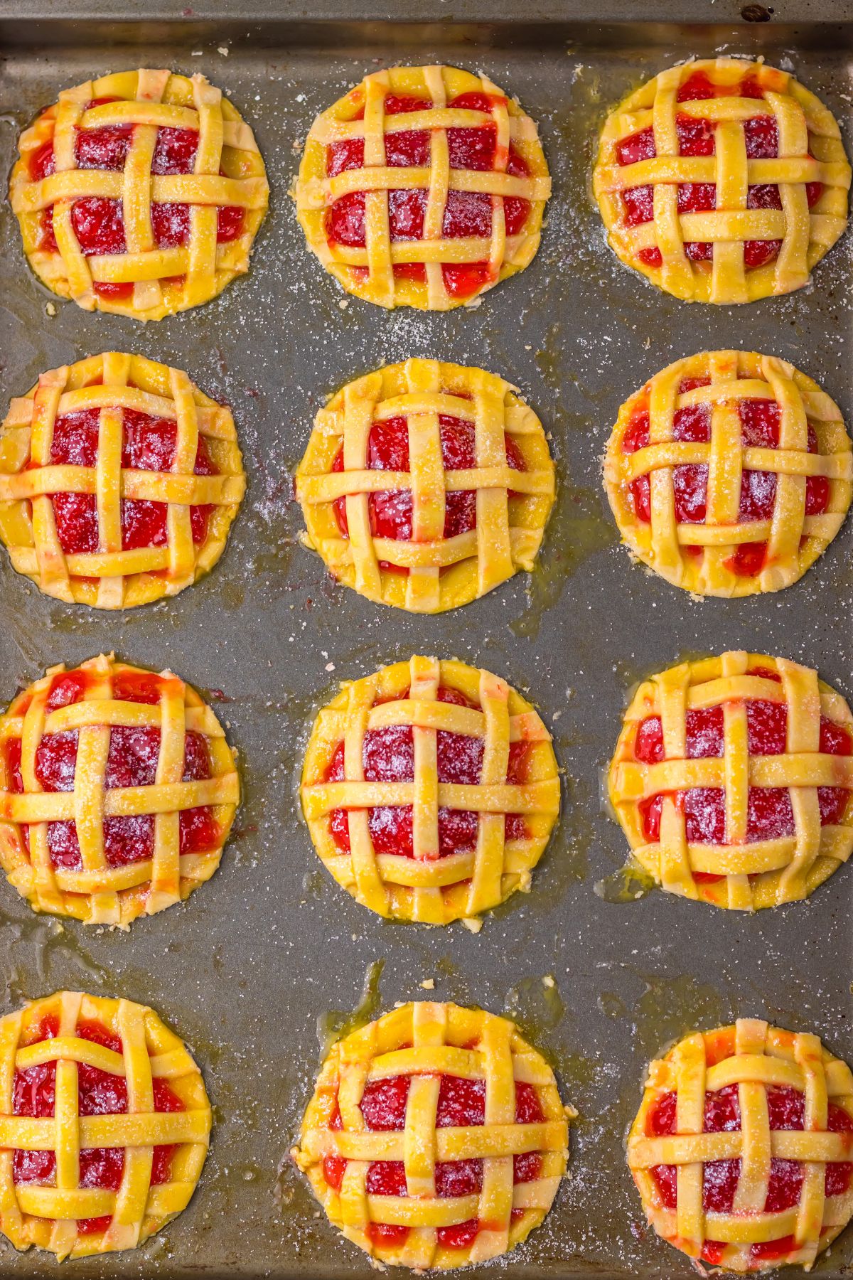 Twelve mini pies with lattice crusts, filled with a red fruit filling, arranged in rows on a baking tray.